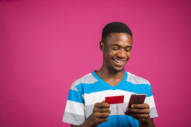 A portrait of an African man shopping online with his phone and credit card against a pink background - 写真・画像