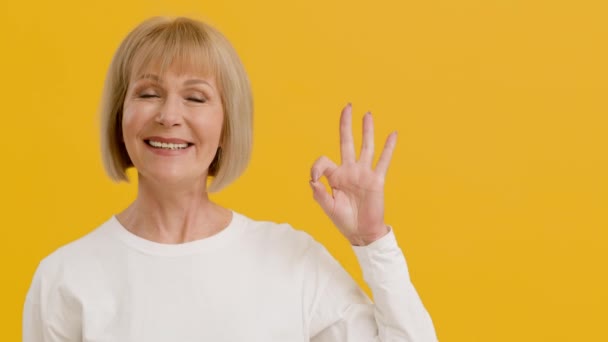 Cheerful senior lady smiling to camera and showing OK gesture, feeling happy, orange background with empty space - Footage, Video