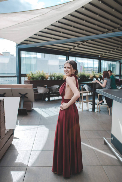 A smiling gorgeous brunette woman in a fancy red dress posing in an outdoor cafe on a sunny day - Photo, image