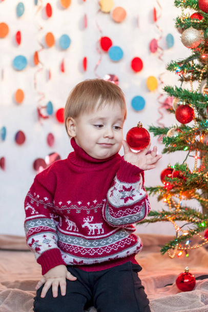 A child is holding a Christmas tree toy while sitting under an elegant Christmas tree. Children's emotions. Christmas and New year. The concept of an advertising banner. - Fotoğraf, Görsel
