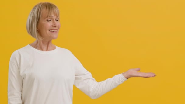 Kijk hier. Positieve volwassen dame strekte haar hand, tonen tekst of product, vrije ruimte, oranje studio achtergrond - Video