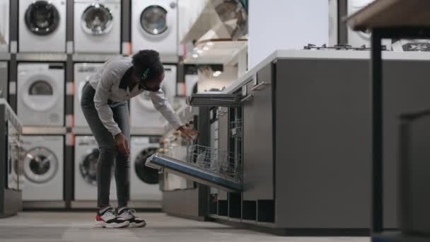 winkelen in huishoudelijke apparaten winkel tijdens pandemie tijd, jonge zwarte vrouw met gezichtsmasker is het bekijken van vaatwasser - Video