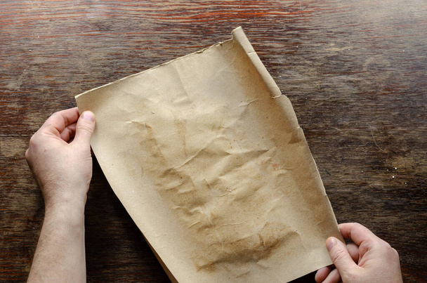 Brown sheet of paper and hands on a wooden table. A man holds a piece of torn brown paper on a scratched wooden surface. Top view, flat lay - Foto, Imagem