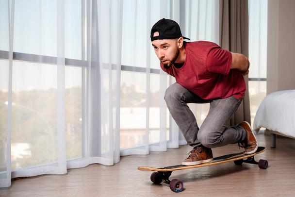 A young man in casual clothes poses on a longboard, imagining that he is driving down the street. Indoor. Concept of quarantine and activity at home. - Foto, Imagen