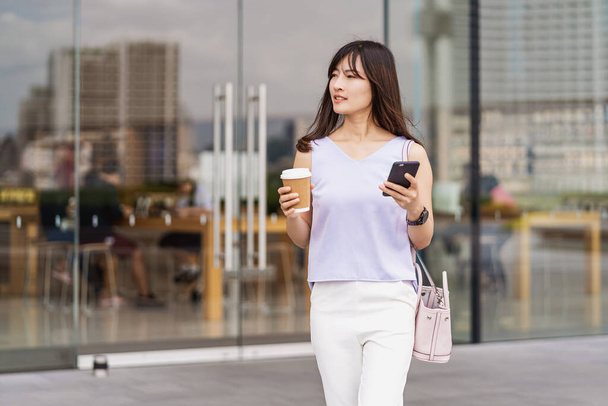 Ázsiai nő sétál a városban, mobilt használ, és egy csésze papírkávét tart a kezében. áruház bevásárló központ, életmód és szabadidő, online vásárlás és készpénz nélküli fizetési koncepció - Fotó, kép