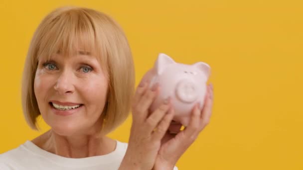 Retirement savings. Cheerful senior lady shaking piggy bank and smiling to camera, orange studio background, slow motion - Footage, Video