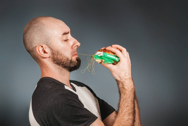 Environmental pollution and food industry. A bald man with a beard holds a hamburger with both hands, from which a filling of thread and debris stretches. Copy space. - Photo, Image