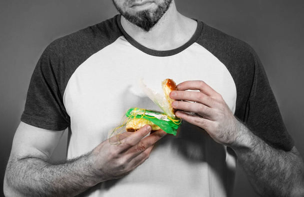 Pollution de l'environnement et industrie alimentaire. Portrait d'un homme à la barbe, qui, perplexe, révèle un hamburger bourré d'ordures. Monochrome. - Photo, image