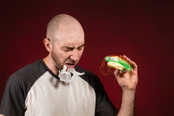 Koncepcja fast food i ekologii. Łysy facet z brodą, trzymający hamburgera i plujący plastikowym nadzieniem. Tło burgundzkie. - Zdjęcie, obraz