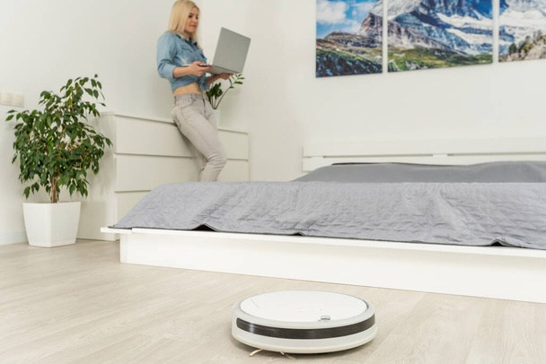 happy woman using laptop while robotic vacuum cleaner washing in living room - Photo, Image