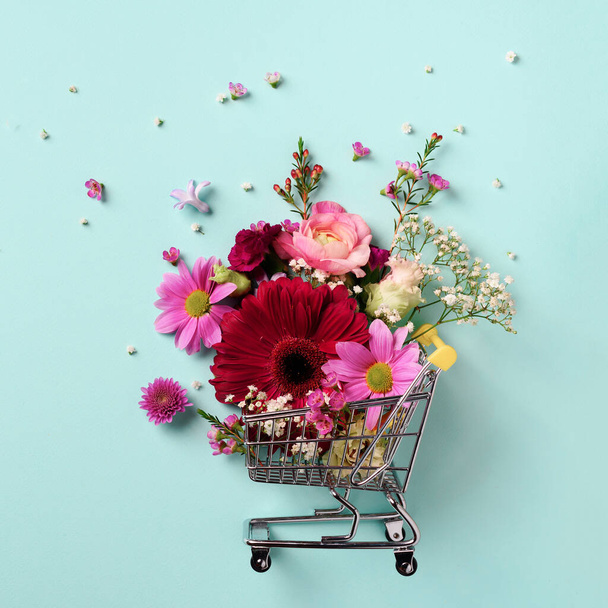 Shopping trolley with flowers on blue punchy pastel background. Banner with copy space. Top view, flat lay. Creative layout. Festive shopping and sale. Gifts for her. - Foto, Bild