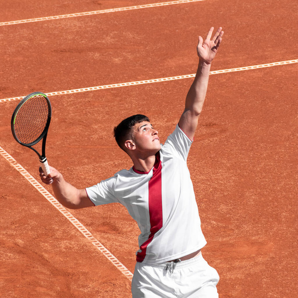 Young male tennis player performs serves ball on clay tennis court at start of game. Cute man athlete in action. Individual, competitive sport - Photo, Image