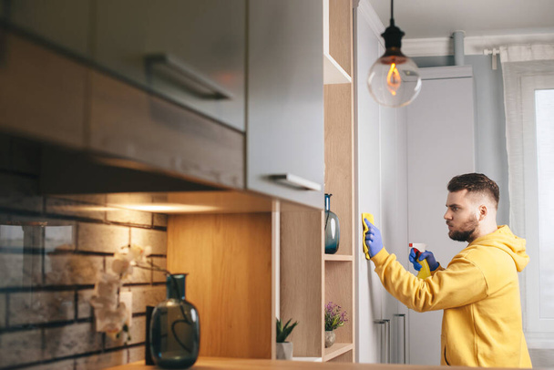 Biały mężczyzna z brodą w żółtym swetrze czyści dom w rękawiczkach i używa sprayu. - Zdjęcie, obraz