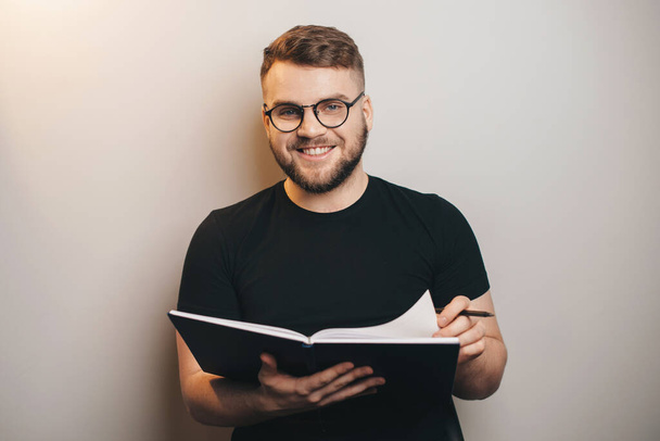 Mann mit Bart lächelt während eines Webinars in die Kamera, hält ein Werbebuch in der Hand und lächelt an einer Studiowand - Foto, Bild
