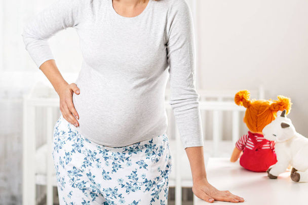 A young pregnant woman is in pain and holds her hand over her stomach, leaning against the dresser. Children's room in the background. The concept of health of a pregnant woman, motherhood. Close up. - Photo, Image