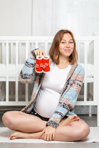 Eine junge schwangere Frau sitzt auf dem Boden und hält lächelnd Booties in der Hand. Konzept von Schwangerschaft und glücklicher Mutterschaft. - Foto, Bild