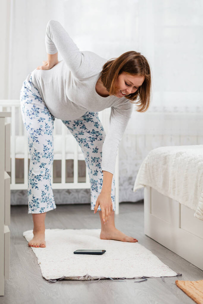 Eine Schwangere kann sich wegen Rückenschmerzen und einem dicken Bauch kaum bücken, um ein am Boden liegendes Smartphone aufzuheben. Konzept der alltäglichen Schwierigkeiten schwangerer Frauen. Seitenansicht. - Foto, Bild