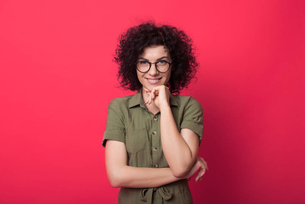 Szép fotó egy kaukázusi göndör hajú nőről, aki szemüveget visel, és a kamera előtt mosolyog a vörös stúdió falán. - Fotó, kép