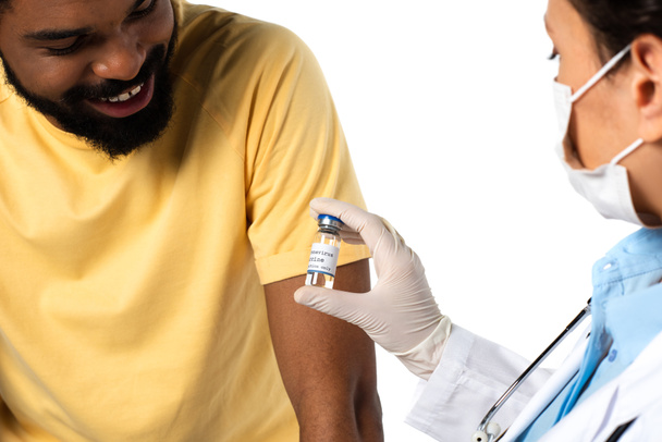 Vaccine in hand of doctor on blurred foreground near smiling african american patient isolated on white  - Photo, Image