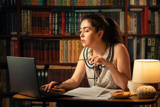Młoda kobieta z okularami w ręku, starannie pracująca przy komputerze, na stole to dokumenty. Wieczór, środowisko domowe.Koncepcja kwarantanny, pracy zdalnej i freelancerstwa. - Zdjęcie, obraz