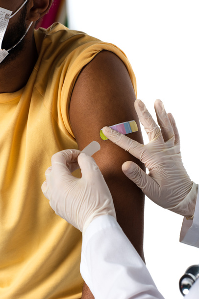 Ausgeschnittene Ansicht eines Arztes in Latexhandschuhen, der Klebepflaster auf den Arm eines afrikanisch-amerikanischen Patienten aufträgt, isoliert auf weiß  - Foto, Bild