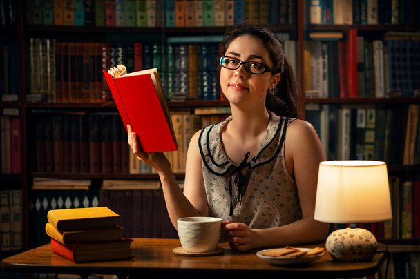 Cuarentena. Una joven mujer caucásica con gafas está leyendo un libro en la mesa, sosteniendo una taza de té. La biblioteca de la casa está en el fondo. Concepto de autoaislamiento, aprendizaje a distancia y trabajo a distancia. - Foto, Imagen