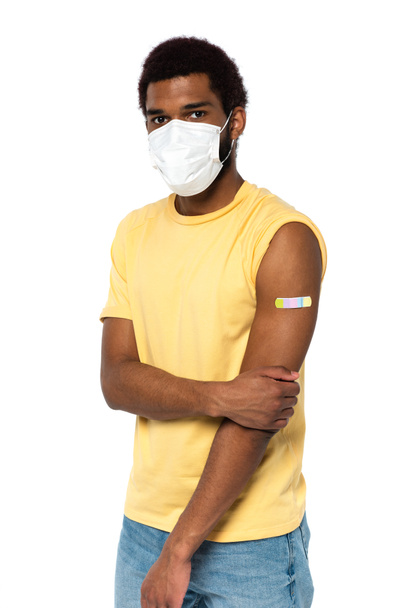 Hombre afroamericano en máscara médica y yeso adhesivo en el brazo mirando a la cámara aislada en blanco  - Foto, Imagen