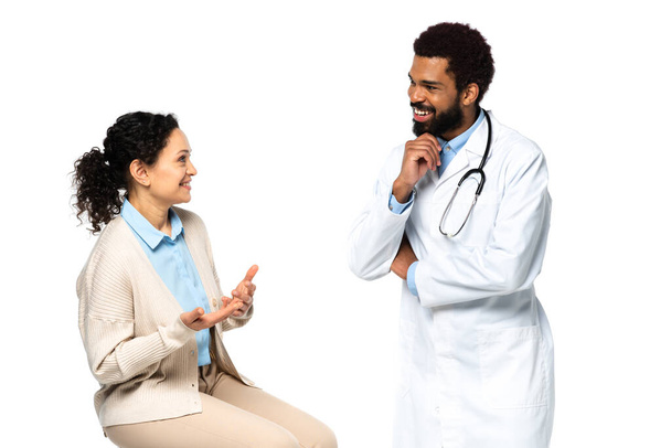 Χαμογελώντας αφροαμερικανός ασθενής μιλώντας με τον γιατρό με λευκό παλτό που απομονώνεται σε λευκό  - Φωτογραφία, εικόνα