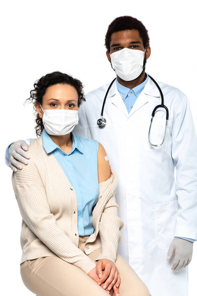 Afrikanisch-amerikanischer Arzt in medizinischer Maske umarmt Patient mit Klebepflaster isoliert auf weißem Grund  - Foto, Bild