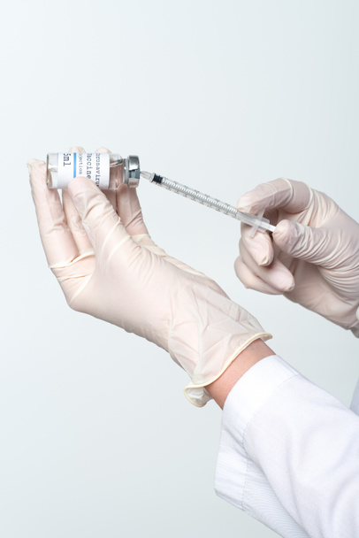 Az orvos levágott nézete latex kesztyűben a szürkén izolált fecskendőben lévő koronavírus vakcina felvétele - Fotó, kép