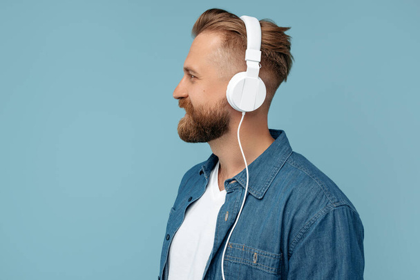 Portrait of bearded blonde hipster man in jeans shirt wearing headphones and looking away over blue background. - Foto, imagen