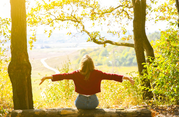 Stylish hipster girl is sitting in forest. Happy woman enjoying autumn nature. Beautiful landscape. Lifestyle travel concept. Dark red sweater. Yellow leaves on trees. View from mountain hill. - Photo, Image
