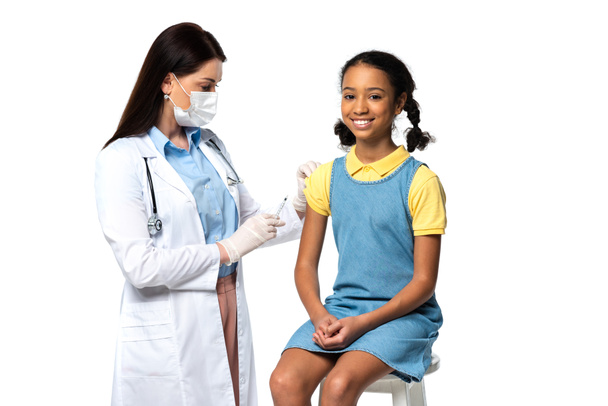Médecin en masque médical tenant la seringue près joyeux enfant afro-américain isolé sur blanc  - Photo, image