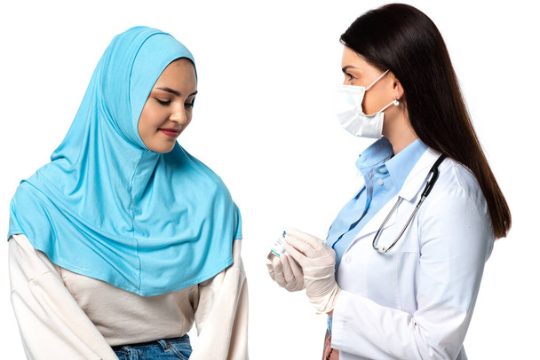 Lekarz w masce medycznej posiadający szczepionkę w pobliżu pacjenta arabskiego izolowanego na białym  - Zdjęcie, obraz