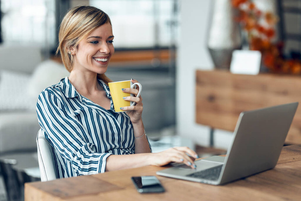 Shot of smiling young business woman working with computer while drinking coffee in the living room at home. - Photo, image