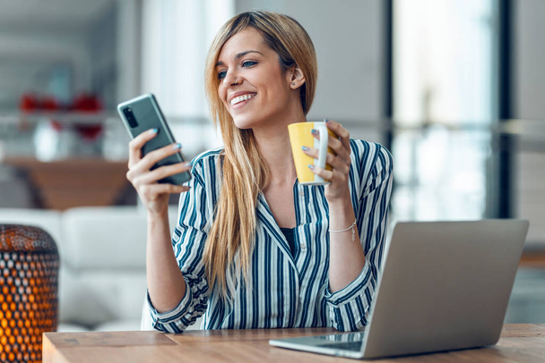 Shot of smiling young woman sending audio message with her smart phone while drinking coffe in the office at home. - Zdjęcie, obraz