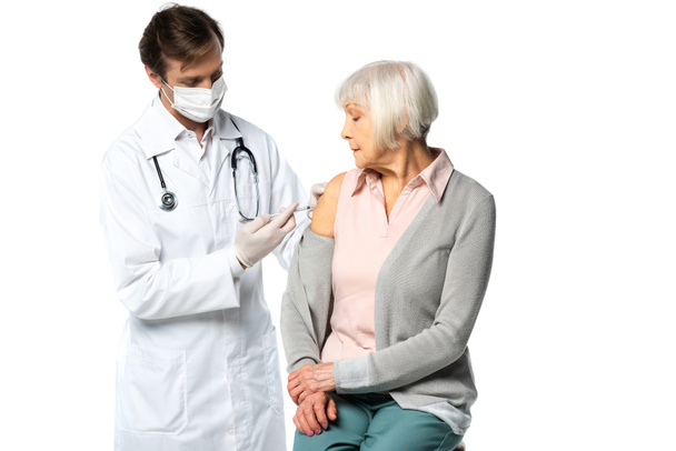 Arzt in medizinischer Maske injiziert Impfstoff an ältere Patientin isoliert auf weiß  - Foto, Bild