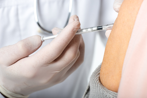 Viljelty näkymä lääkäristä lateksikäsineissä tekemässä injektiota rokotteella iäkkäämmällä potilaalla, eristettynä valkoisella  - Valokuva, kuva