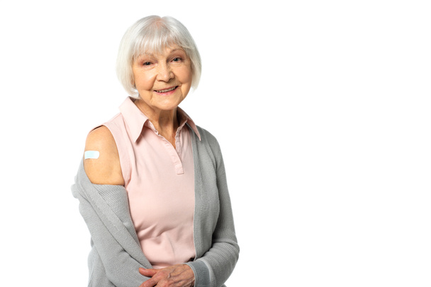 Mujer anciana positiva con yeso adhesivo en el brazo mirando a la cámara aislada en blanco  - Foto, Imagen