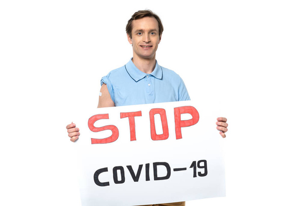Smiling man with adhesive plaster on arm holding placard with stop covid- 2019 lettering isolated on white  - 写真・画像