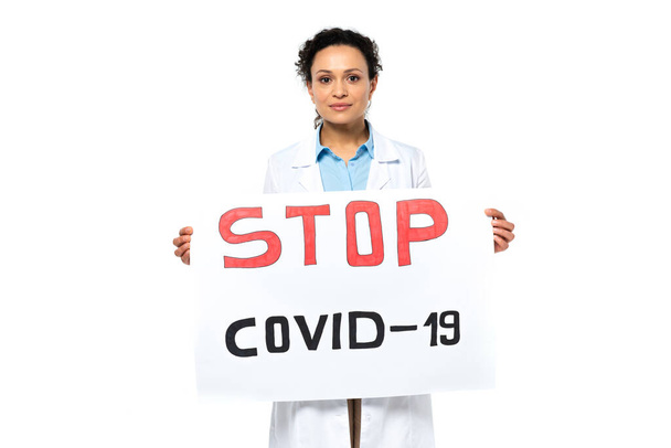 Afrykański amerykański lekarz trzymający plakietkę z przystankiem covid-2019 literowanie i patrząc na kamerę odizolowaną na białym  - Zdjęcie, obraz