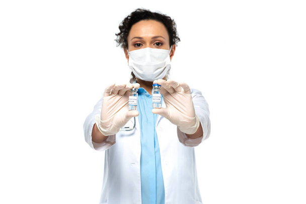 Frascos con vacuna en manos del médico afroamericano en máscara médica sobre fondo borroso aislado sobre blanco  - Foto, Imagen