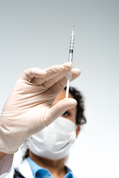Χαμηλή γωνία όψης της σύριγγας με εμβόλιο στο χέρι του θολωμένου Αφροαμερικανού γιατρού με ιατρική μάσκα που απομονώνεται στο γκρι  - Φωτογραφία, εικόνα