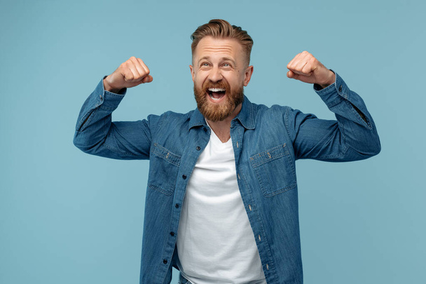 Excited bearded hipster blonde man in jeans shirt celebrate success over blue background. - Φωτογραφία, εικόνα