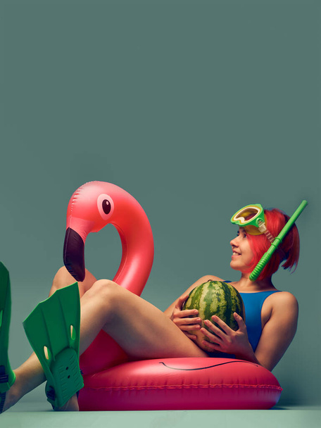 Girl sitting on big inflatable flamingo, ready to swim, blue studio background with empty space - Foto, Imagem
