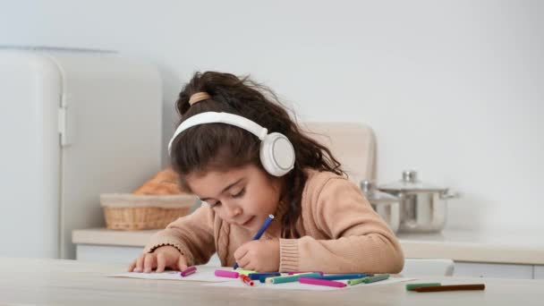 Pequeña niña dibujando usando auriculares inalámbricos sentada en la cocina interior - Imágenes, Vídeo