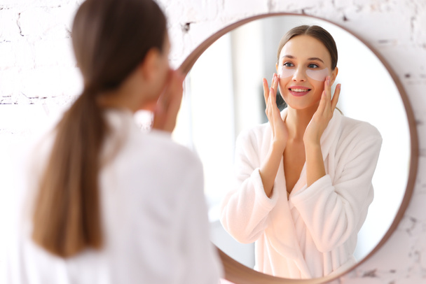 Szczęśliwa młoda kobieta w białym szlafroku nakłada plastry nawilżające pod oczami przed lustrem w łazience - Zdjęcie, obraz