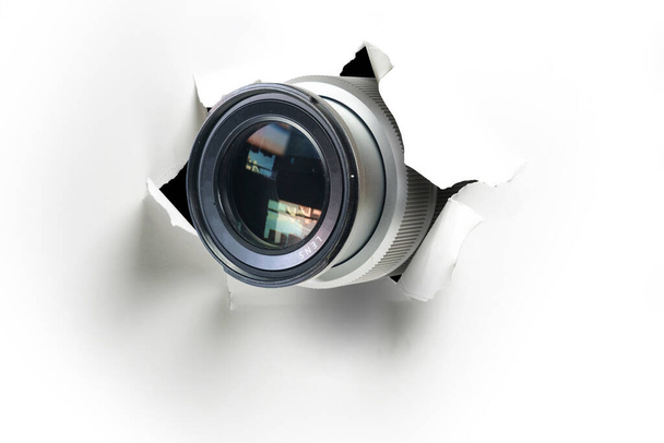 Concept of paparazzi or hidden camera, camera lens looks out through a hole in white paper wall - Photo, Image