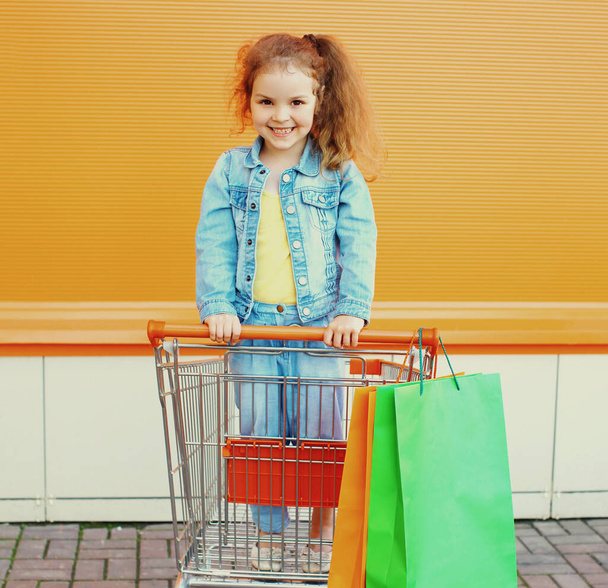 Portrait of little girl child with trolley cart and shopping bags wearing a denim clothes on a city street - 写真・画像