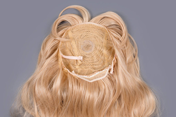 Blonde Perücke aus nächster Nähe auf grauem Hintergrund. Goldene Echthaare weben sich, Verlängerungen und Perücken. Schönheitskonzept für Frauen - Foto, Bild